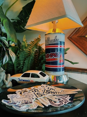 A decorative table setting featuring a lamp with a base resembling an Atlanta Braves can. Surrounding the lamp are a collection of stickers, a toy car marked 'Joe Chitwood's Thrill', a small carved frog figure, and lush green plants in the background. The scene is warmly lit, with a focus on vintage and nostalgic elements.