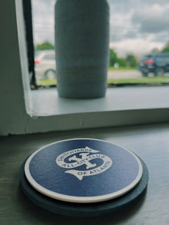 A round blue coaster with the text 'Switchyards All-City Club of Atlanta' sits on a windowsill. In the background, outside the window, there is a blurred view of cars on a road and trees under a cloudy sky. A cylindrical object, possibly a vase, is also on the windowsill.