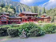 A traditional Japanese-style temple with red and black roofing is set against a backdrop of lush green mountains and tall trees. In the foreground, there is a garden with neatly trimmed bushes, and a stone statue of a seated figure adorned with a small orange garland. The sky is partly cloudy, and the overall scene exudes tranquility.