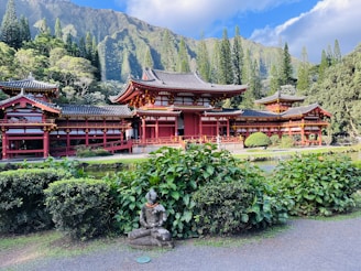 A traditional Japanese-style temple with red and black roofing is set against a backdrop of lush green mountains and tall trees. In the foreground, there is a garden with neatly trimmed bushes, and a stone statue of a seated figure adorned with a small orange garland. The sky is partly cloudy, and the overall scene exudes tranquility.
