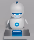 A small, white humanoid robot with blue accents, including eyes, mouth, and a circular badge with the letters 'AI' on its chest, is positioned in front of a blue laptop on a metallic surface. The robot has a simple, smooth design with two cylindrical arms and a small antenna on top.