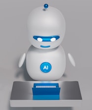 A small, white humanoid robot with blue accents, including eyes, mouth, and a circular badge with the letters 'AI' on its chest, is positioned in front of a blue laptop on a metallic surface. The robot has a simple, smooth design with two cylindrical arms and a small antenna on top.