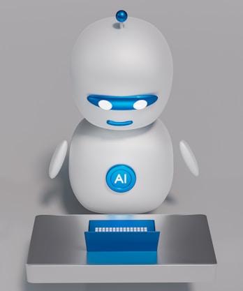 A small, white humanoid robot with blue accents, including eyes, mouth, and a circular badge with the letters 'AI' on its chest, is positioned in front of a blue laptop on a metallic surface. The robot has a simple, smooth design with two cylindrical arms and a small antenna on top.