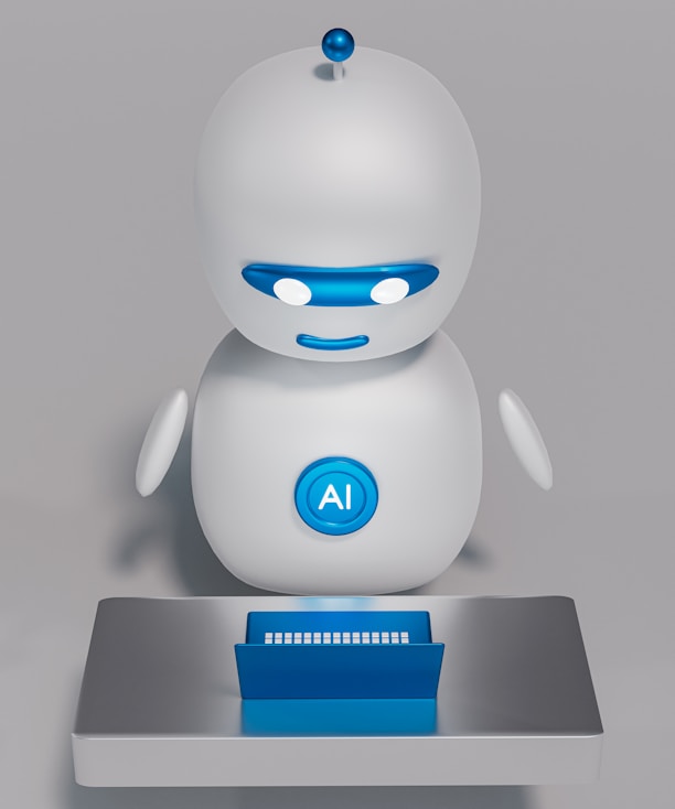 A small, white humanoid robot with blue accents, including eyes, mouth, and a circular badge with the letters 'AI' on its chest, is positioned in front of a blue laptop on a metallic surface. The robot has a simple, smooth design with two cylindrical arms and a small antenna on top