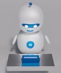 A small, white humanoid robot with blue accents, including eyes, mouth, and a circular badge with the letters 'AI' on its chest, is positioned in front of a blue laptop on a metallic surface. The robot has a simple, smooth design with two cylindrical arms and a small antenna on top.
