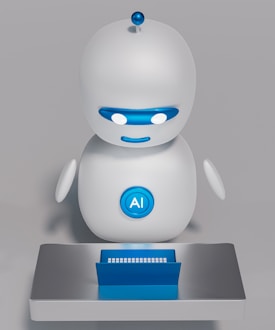 A small, white humanoid robot with blue accents, including eyes, mouth, and a circular badge with the letters 'AI' on its chest, is positioned in front of a blue laptop on a metallic surface. The robot has a simple, smooth design with two cylindrical arms and a small antenna on top.