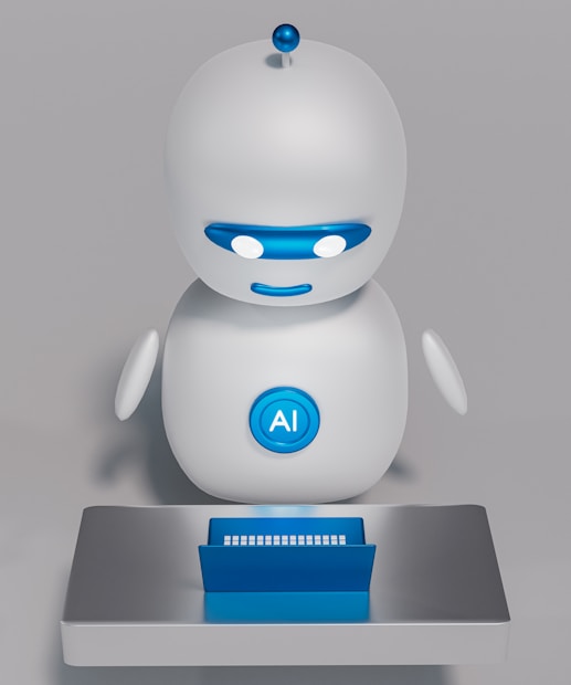 A small, white humanoid robot with blue accents, including eyes, mouth, and a circular badge with the letters 'AI' on its chest, is positioned in front of a blue laptop on a metallic surface. The robot has a simple, smooth design with two cylindrical arms and a small antenna on top.
