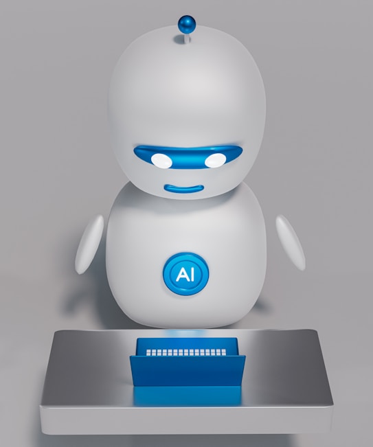 A small, white humanoid robot with blue accents, including eyes, mouth, and a circular badge with the letters 'AI' on its chest, is positioned in front of a blue laptop on a metallic surface. The robot has a simple, smooth design with two cylindrical arms and a small antenna on top.
