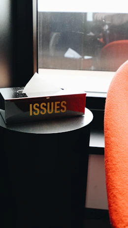 A black tissue box with the word 'ISSUES' in bold, yellow letters sits on a round black side table by the window. A single white tissue is protruding from the box. Next to the table, there is an orange-red upholstered chair partially visible.