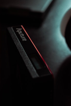 A close-up view of a rectangular electronic device with the brand name 'Aputure' visible on its side. The lighting is dim, creating a dramatic and moody effect. The device appears to have a small digital display and a sleek, modern design.
