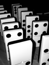A collection of standing dominoes arranged closely together on a wooden surface. Each domino is white with a black dot pattern, indicating different values. The lighting casts sharp shadows and highlights the glossy finish of the dominoes, creating a dramatic contrast.