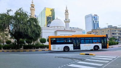 A cityscape featuring a large modern bus traveling on a road. The background includes an ornate building with domes and minarets, possibly a mosque, alongside high-rise buildings with reflective glass windows. Greenery borders the scene, with a large tree on the left.