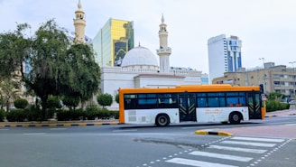 A cityscape featuring a large modern bus traveling on a road. The background includes an ornate building with domes and minarets, possibly a mosque, alongside high-rise buildings with reflective glass windows. Greenery borders the scene, with a large tree on the left.