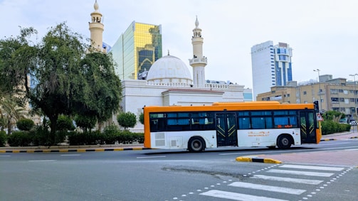 A cityscape featuring a large modern bus traveling on a road. The background includes an ornate building with domes and minarets, possibly a mosque, alongside high-rise buildings with reflective glass windows. Greenery borders the scene, with a large tree on the left.