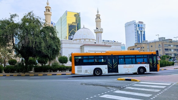 A cityscape featuring a large modern bus traveling on a road. The background includes an ornate building with domes and minarets, possibly a mosque, alongside high-rise buildings with reflective glass windows. Greenery borders the scene, with a large tree on the left.