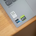 A close-up view of a laptop corner displaying various stickers, including a QR code, Dolby Atmos logo, Intel Evo label, and NVIDIA GeForce RTX badge. The surface is silver, and the laptop is positioned on a wooden desk.