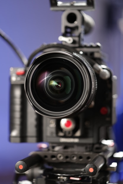 A professional camera with a large lens is positioned on a tripod. The focus is on the lens, which is prominent in the foreground, while the camera body is slightly blurred. The background is a gradient of soft blue tones, creating a contrasting backdrop for the dark, sleek equipment.
