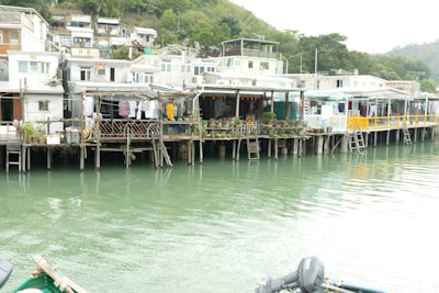 A series of stilt houses line a waterfront, with clothes hanging on lines and an array of potted plants on the balconies. The houses are built closely together and exhibit a rustic appearance, with a hill filled with greenery in the background.