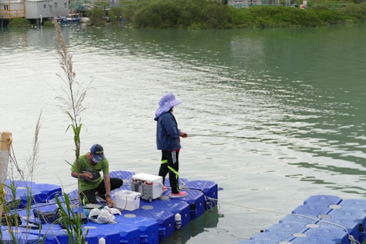 Two people are engaging in fishing activities on a floating platform by a riverside. One person is standing with a fishing rod while the other is sitting and attending to fishing equipment. The water is calm, and the banks are lined with greenery. The platform is made of blue interconnected blocks.