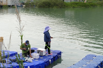 Two people are engaging in fishing activities on a floating platform by a riverside. One person is standing with a fishing rod while the other is sitting and attending to fishing equipment. The water is calm, and the banks are lined with greenery. The platform is made of blue interconnected blocks.