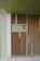 A modern, minimalist entrance with vertical wooden slats and a soft green frame dominates the image. A circular surveillance camera is mounted above the doorway. There are control panels and a number '10' featured beside the entrance.