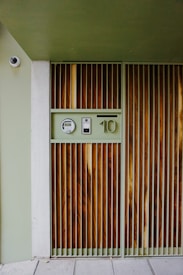 A modern, minimalist entrance with vertical wooden slats and a soft green frame dominates the image. A circular surveillance camera is mounted above the doorway. There are control panels and a number '10' featured beside the entrance.