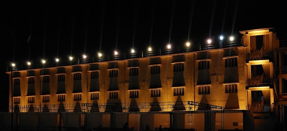 A large building illuminated by a series of bright lights along the upper edge, casting shadows downward. The architecture features multiple windows with strong geometric patterns formed by the interplay of light and shadow. The building appears to have multiple stories and a flat roof, with an outdoor staircase on the right side.