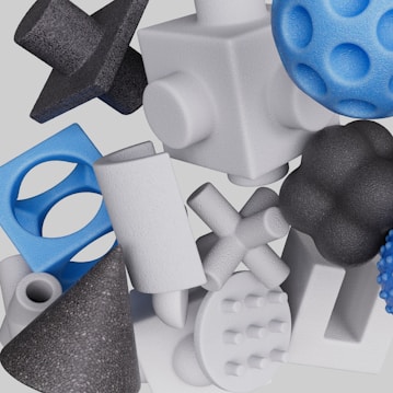 A collection of various 3D geometric shapes, including cubes, spheres, crosses, cones, and cylinders. The textures are rough and the objects are arranged in a seemingly random overlapping fashion. The color palette consists mostly of whites, blues, and blacks.