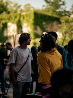 A group of individuals stands in an outdoor setting illuminated by soft, natural lighting. Two people, one wearing a white shirt and another in a yellow hoodie, are engaged in conversation. The background features a building adorned with lush green ivy, suggesting a relaxed, natural environment. Other people are blurred in the background, creating a lively, social scene.
