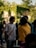 A group of individuals stands in an outdoor setting illuminated by soft, natural lighting. Two people, one wearing a white shirt and another in a yellow hoodie, are engaged in conversation. The background features a building adorned with lush green ivy, suggesting a relaxed, natural environment. Other people are blurred in the background, creating a lively, social scene.