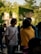 A group of individuals stands in an outdoor setting illuminated by soft, natural lighting. Two people, one wearing a white shirt and another in a yellow hoodie, are engaged in conversation. The background features a building adorned with lush green ivy, suggesting a relaxed, natural environment. Other people are blurred in the background, creating a lively, social scene.
