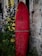 A vibrant red surfboard is propped up against a textured stone wall. The surfboard features a small design or logo near the top and appears weathered with white markings. Lush green foliage and purple flowers surround the base of the surfboard, adding a natural element to the urban setting.