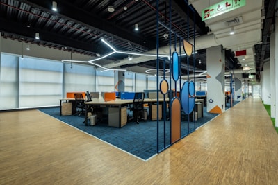A modern office space with an open-plan layout features workstations separated by colorful dividers. It has a combination of wooden and carpeted flooring, and modern ceiling lighting. The walls are adorned with geometric patterns. An accessible exit sign is visible on the ceiling.