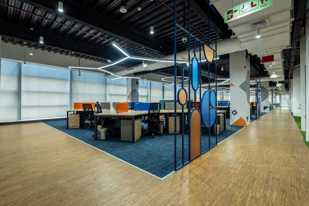 A modern office space with an open-plan layout features workstations separated by colorful dividers. It has a combination of wooden and carpeted flooring, and modern ceiling lighting. The walls are adorned with geometric patterns. An accessible exit sign is visible on the ceiling.