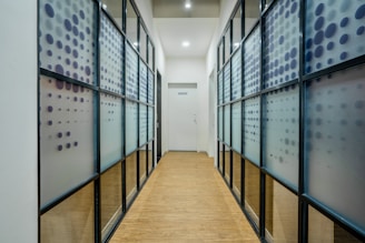 A narrow corridor with wooden flooring is lined with frosted glass walls featuring a pattern of blue and purple dots. The ceiling is white with recessed lighting, and there's a door at the end of the hallway.