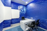 A modern office space features a white desk with two black chairs. The walls are covered with blue wallpaper adorned with digital circuit patterns and a globe, accompanied by a cartoon robot. The room has a futuristic design with a focus on a technological theme.