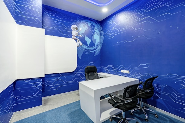 A modern office space features a white desk with two black chairs. The walls are covered with blue wallpaper adorned with digital circuit patterns and a globe, accompanied by a cartoon robot. The room has a futuristic design with a focus on a technological theme.