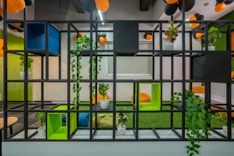 A modern interior space featuring a black grid shelving unit adorned with various potted plants. In the background, there are bright orange and black balloons hanging from the ceiling. The walls and floor incorporate vibrant green and blue accents, creating a lively and colorful atmosphere. This setting includes minimalist furniture such as round tables and bean bags, adding to a cozy and inviting environment.