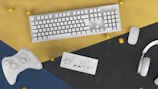 A white keyboard, mouse, game controller, Nintendo Switch, and headphones are arranged on a colored geometric surface with gold accents.