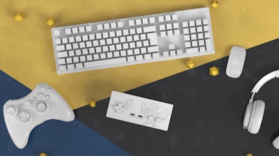 A white keyboard, mouse, game controller, Nintendo Switch, and headphones are arranged on a colored geometric surface with gold accents.