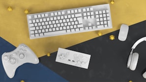 A white keyboard, mouse, game controller, Nintendo Switch, and headphones are arranged on a colored geometric surface with gold accents.