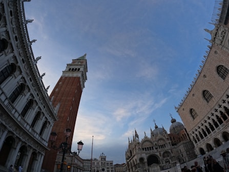 An architectural scene featuring an ornate historical building with a tall, narrow bell tower and decorative stonework on the walls. The sky is clear with a few wispy clouds, and the lighting suggests either late afternoon or early evening.