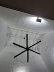 A large black ceiling fan is mounted on a high ceiling with intricate white geometric patterns. Soft lighting creates shadows on the ceiling, enhancing its texture. A series of small string lights are also visible, adding a decorative element.