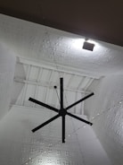 A large black ceiling fan is mounted on a high ceiling with intricate white geometric patterns. Soft lighting creates shadows on the ceiling, enhancing its texture. A series of small string lights are also visible, adding a decorative element.