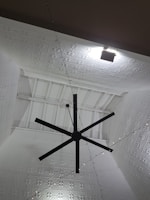 A large black ceiling fan is mounted on a high ceiling with intricate white geometric patterns. Soft lighting creates shadows on the ceiling, enhancing its texture. A series of small string lights are also visible, adding a decorative element.