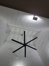 A large black ceiling fan is mounted on a high ceiling with intricate white geometric patterns. Soft lighting creates shadows on the ceiling, enhancing its texture. A series of small string lights are also visible, adding a decorative element.