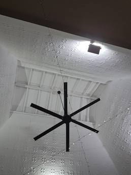 A large black ceiling fan is mounted on a high ceiling with intricate white geometric patterns. Soft lighting creates shadows on the ceiling, enhancing its texture. A series of small string lights are also visible, adding a decorative element.