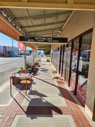 A sidewalk cafe with outdoor seating, featuring tables and chairs along a paved walkway with a canopy cover. A sign reads 'Bottom Cafe: Pizza, Coffee, Take Away'. The street is quiet with buildings visible in the background. Sunlight casts long shadows on the ground.
