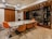 A modern dining room with a sleek marble table surrounded by six orange leather chairs. The room features a contemporary design with wooden paneling on the ceiling, built-in shelves displaying decor pieces, and wood blinds covering the windows. The lighting is warm, enhancing the elegant atmosphere.