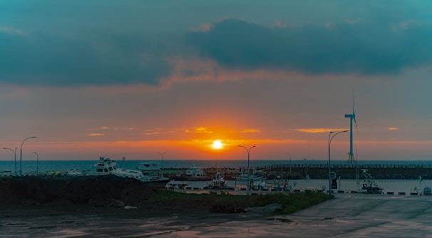 A serene coastal harbor with fishing boats and wind turbines in the background at sunset.
